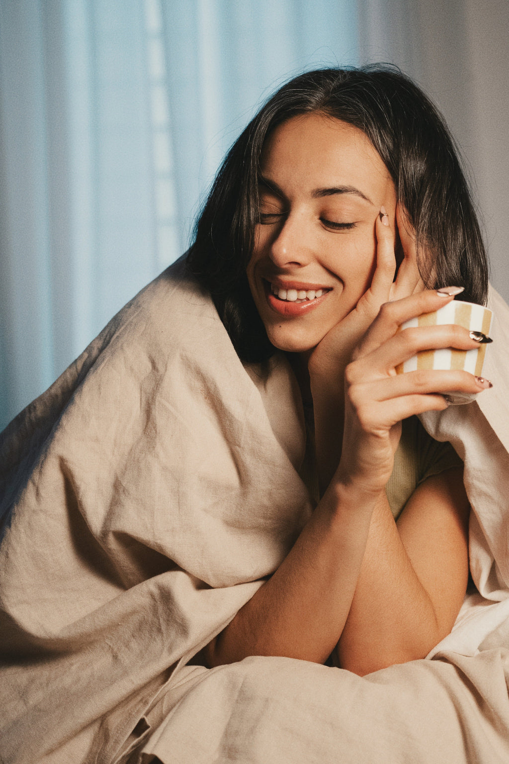 Woman wrapped in a beige blanket holding a mug, smiling.